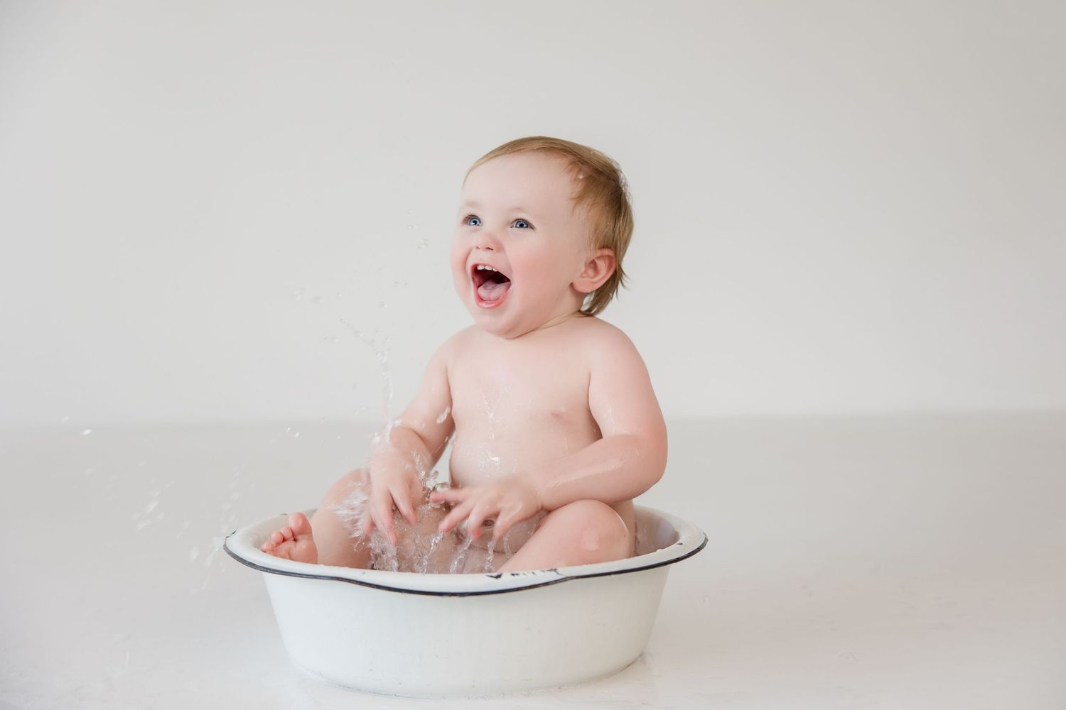 Happy baby sitting in white basin against light background.
