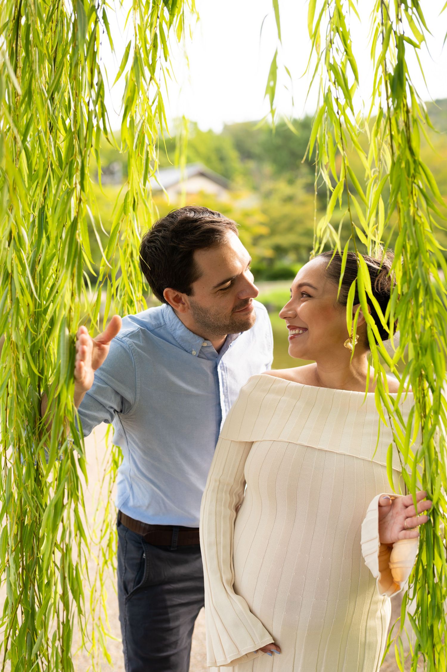 Smiling pregnant couple gazes at each other under cascading willow tree branches in a park.