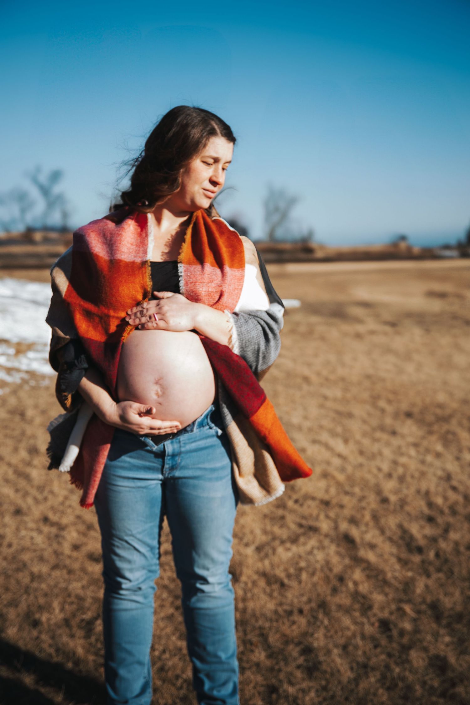 An expectant mother in jeans and orange wrap stands in a field during winter.