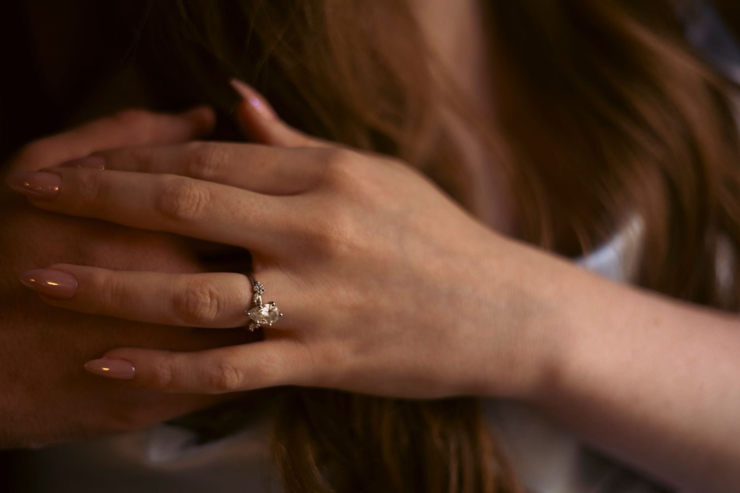 A close-up of a hand with an engagement ring set against soft brown hair in warm lighting.