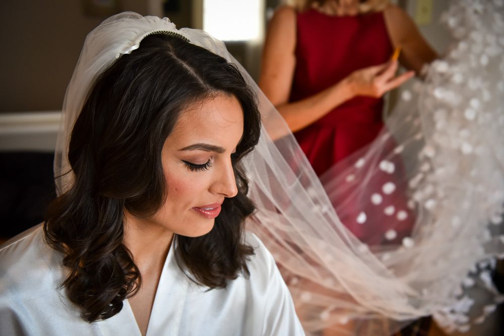 A bride wearing a white robe and delicate veil smiles while getting ready on her wedding day.