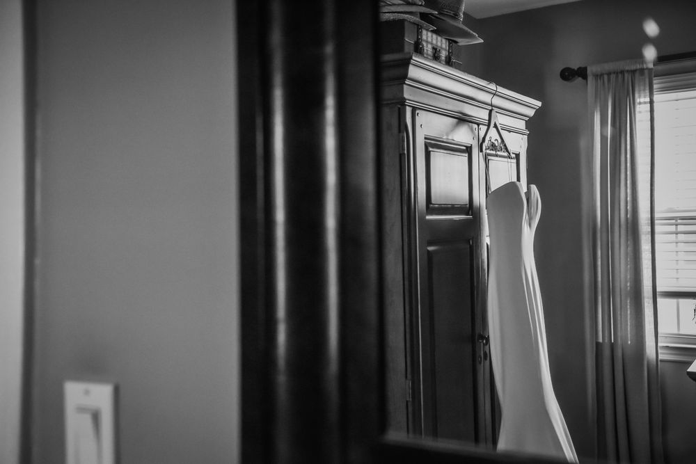A white wedding dress hangs elegantly beside a dark wooden armoire in a black and white photograph.