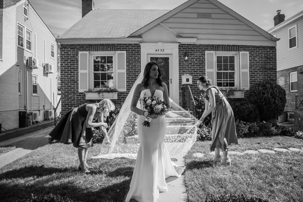 Black and white photo of a bride in wedding dress and helpers adjusting veil on residential home walkway.