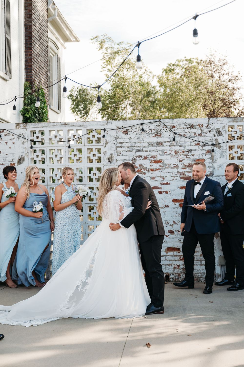 bride & groom kiss during ceremony while bridesmaids & groomsmen smile outside ceremony space at Sister Hill venue by midwest wedding photog