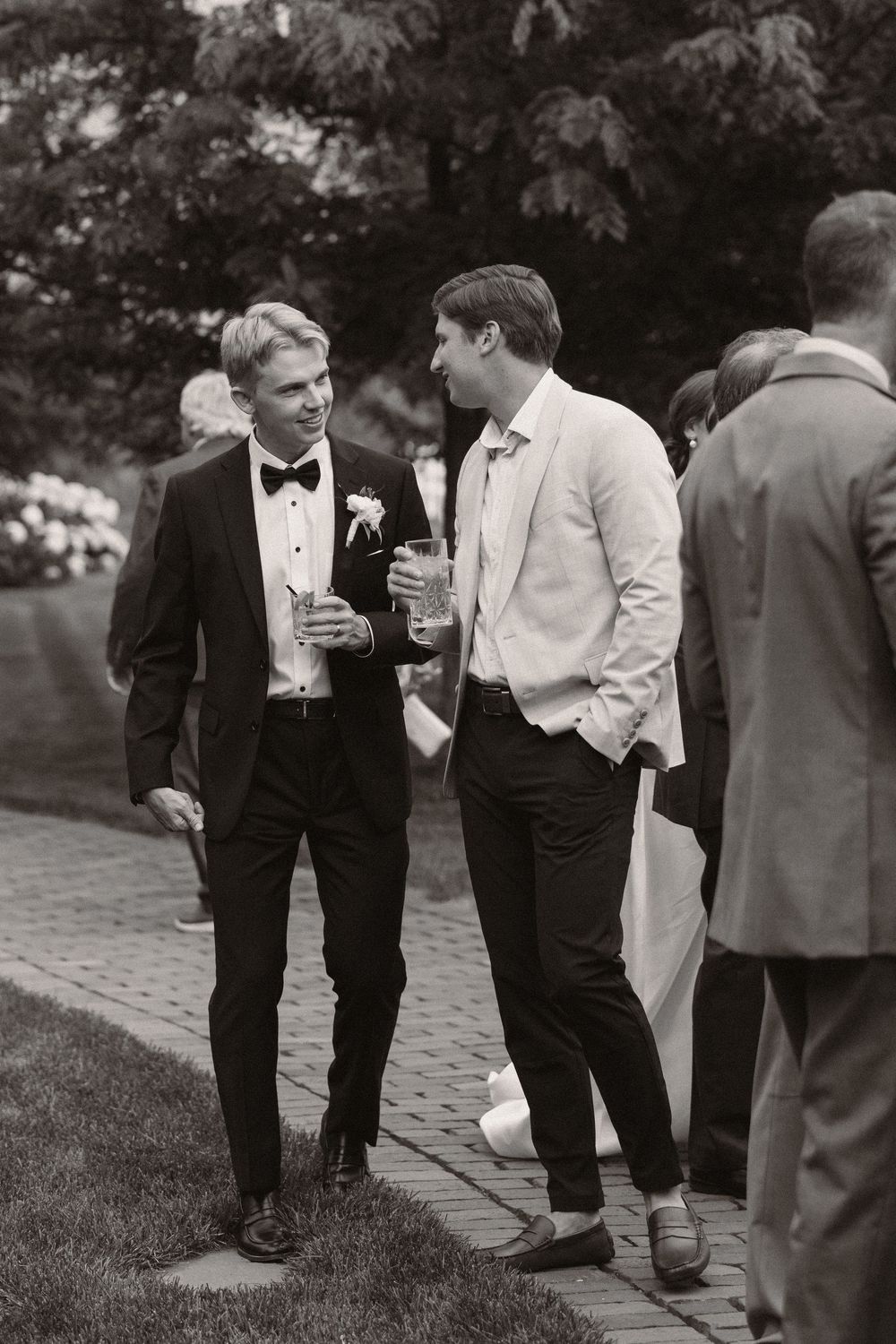 Black and white photo of guests in formal attire socializing on brick pathway.