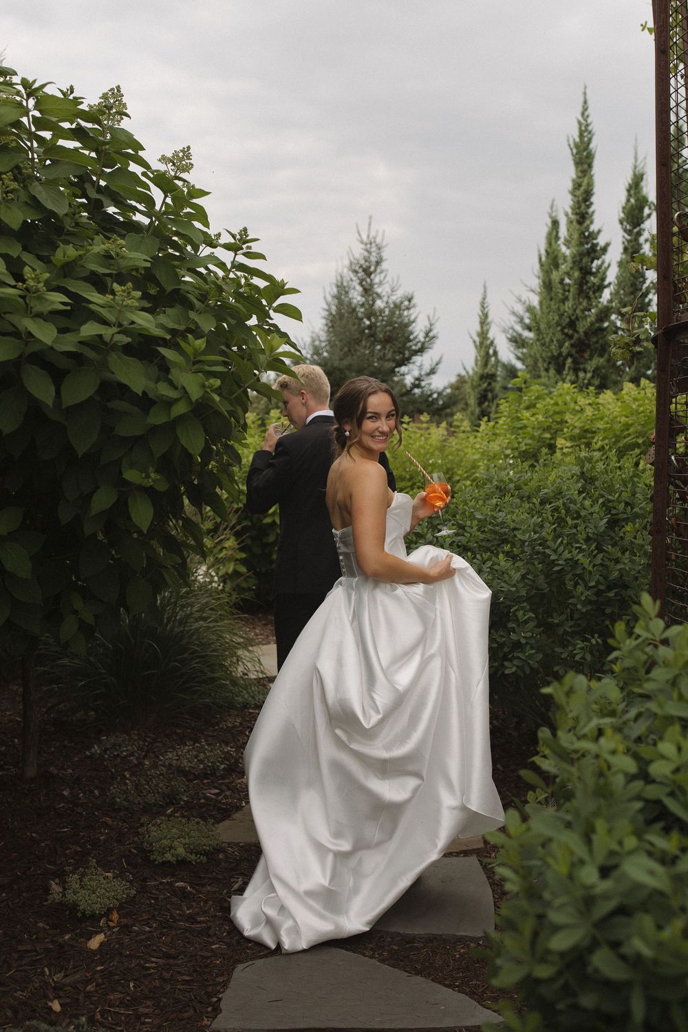 A bride in a white wedding dress sits on a stone bench surrounded by lush greenery in a garden setting.