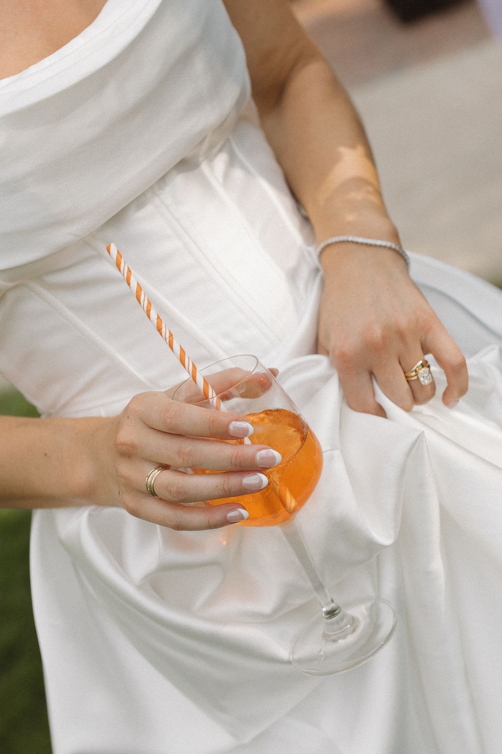 Close-up of hands holding an orange cocktail with a striped straw against a white wedding dress.