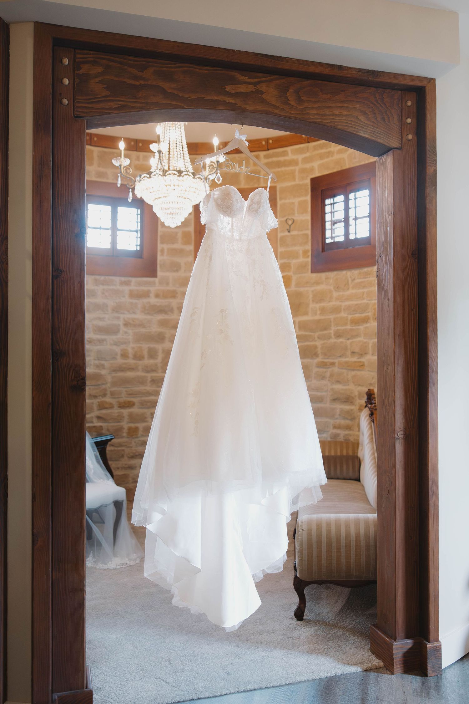 A white wedding dress hangs elegantly suspended in a rustic stone room with dark wood beams and a crystal chandelier.