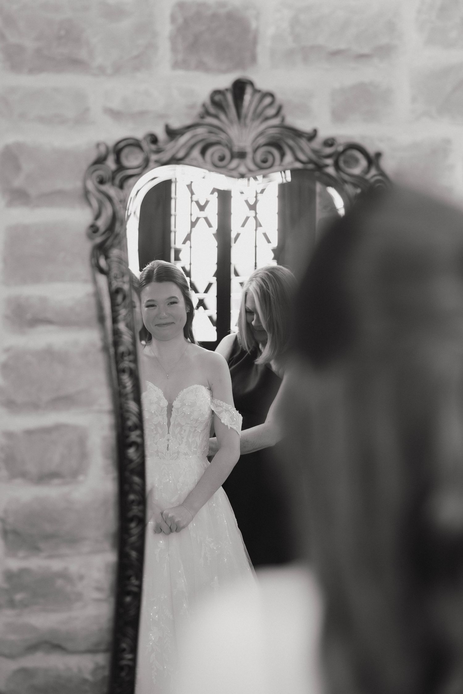 Black and white reflection in ornate mirror shows elegant bride in strapless wedding gown getting ready on wedding day.