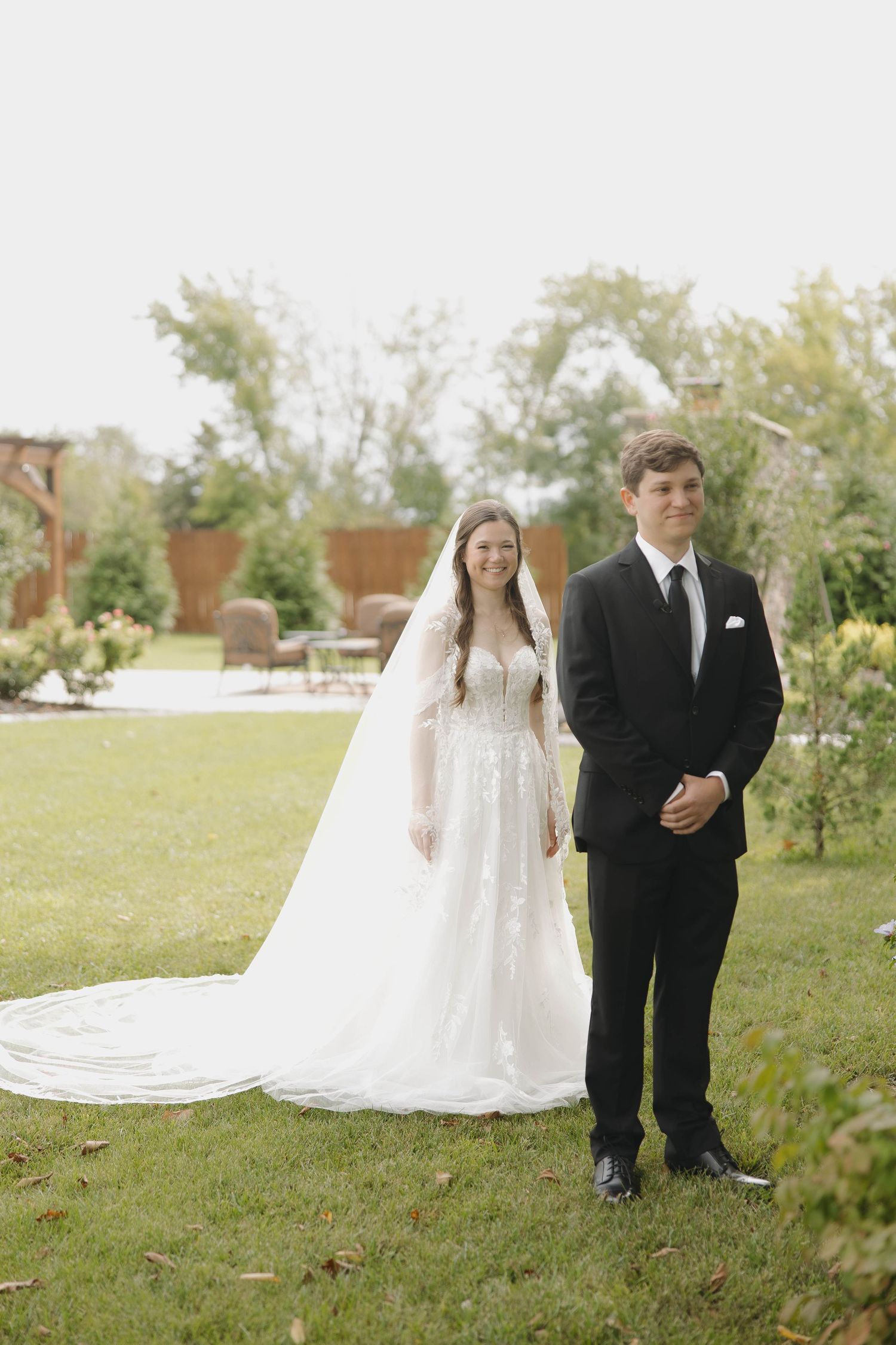 A wedding couple stands together on a grassy lawn, with the bride's long white veil flowing in the breeze.