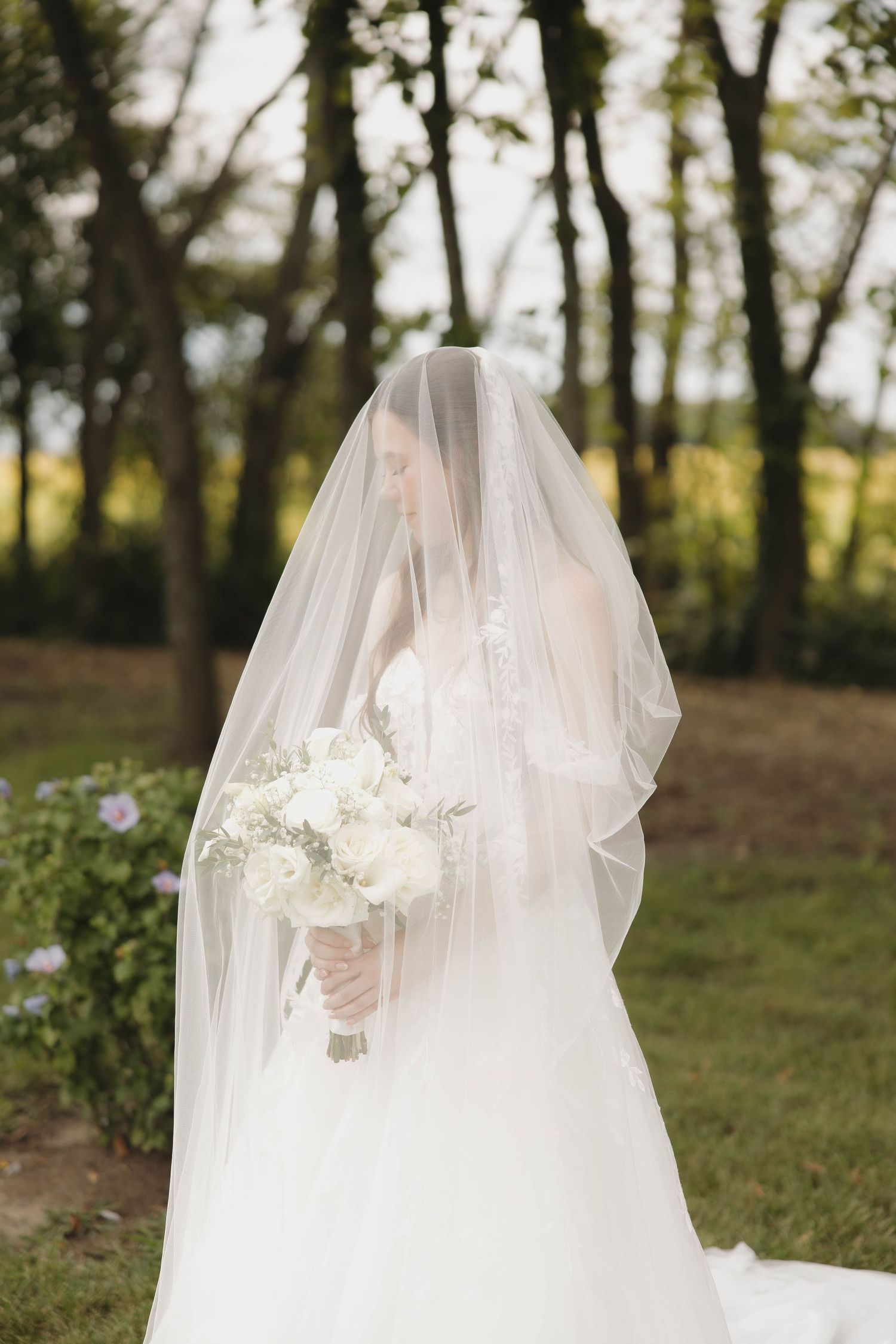 A wedding dress with flowing veil and white bouquet photographed in a dreamy outdoor wooded setting.