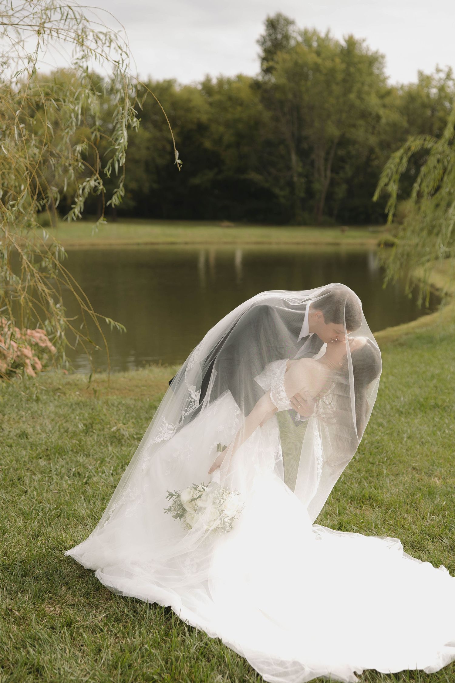 A bride in a flowing white wedding gown and veil poses gracefully near a serene lake surrounded by willow trees.