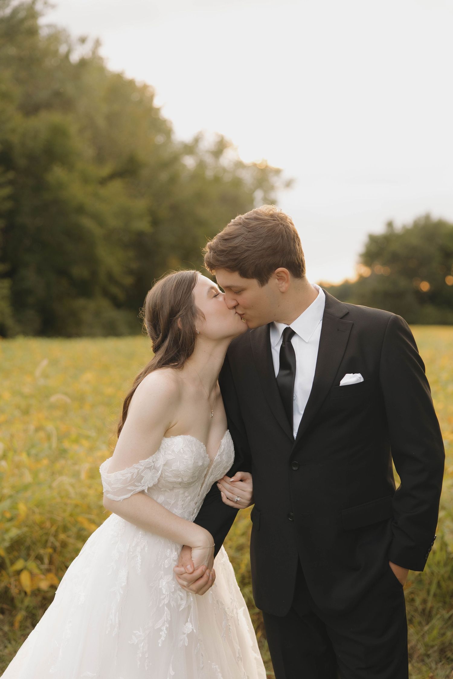 A couple shares romantic sunset kisses in a golden wildflower field while wearing formal wedding attire.