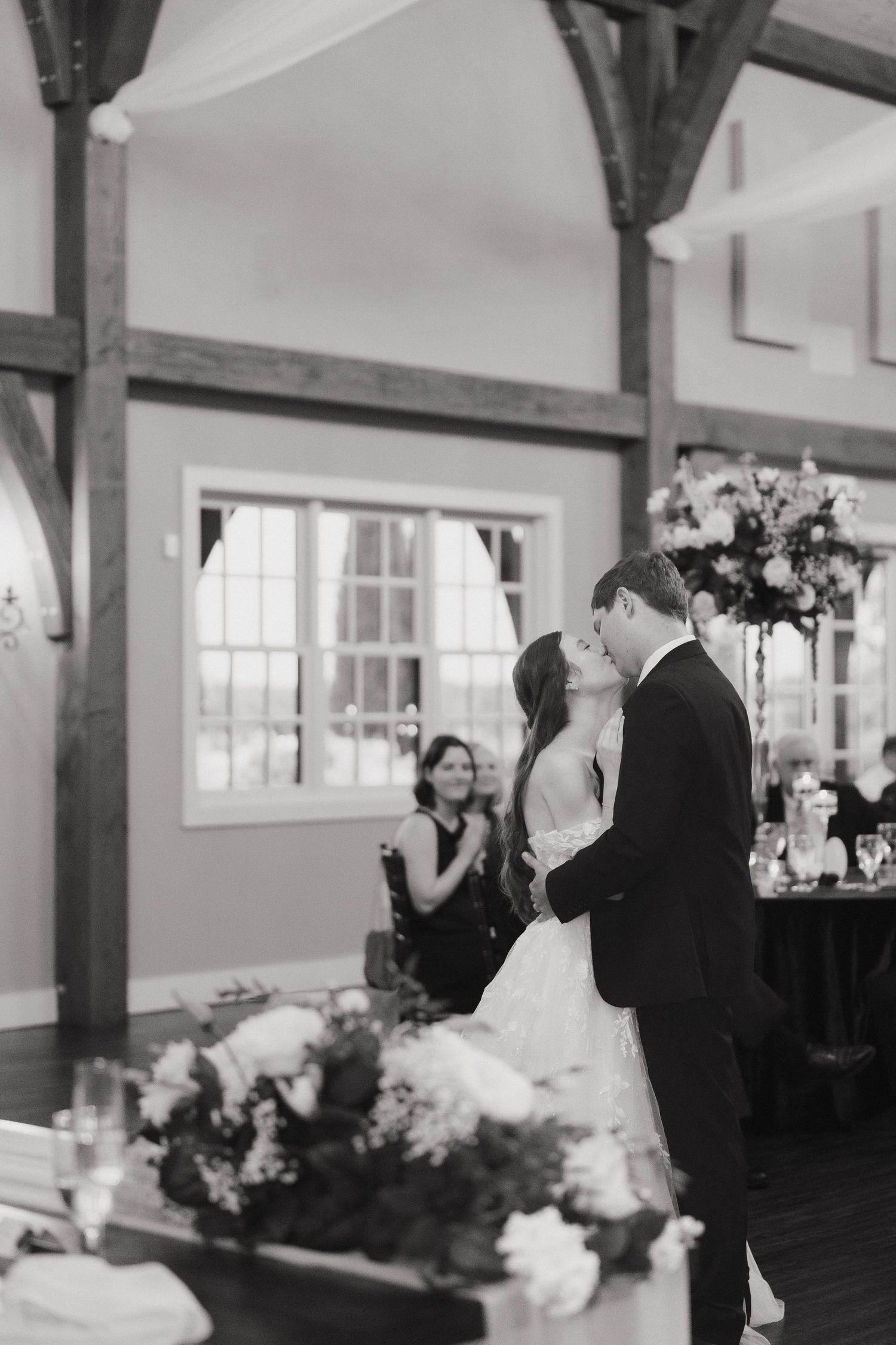 A couple dances in a rustic indoor venue with exposed wooden beams and natural light.