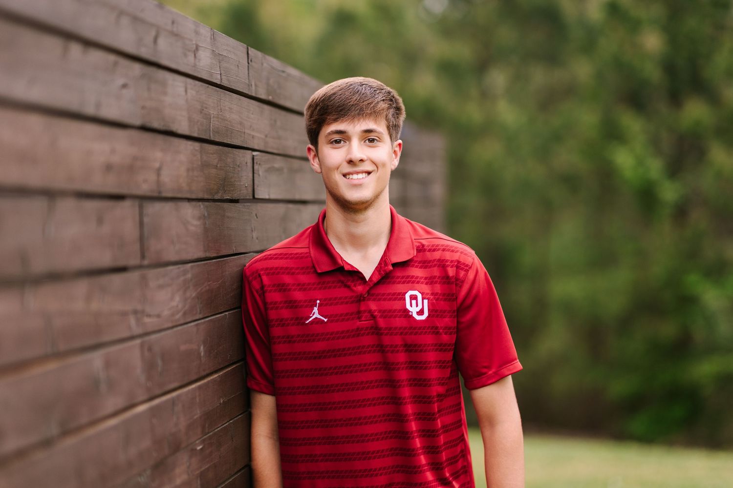 Person wearing a red Jordan Oklahoma University shirt poses outdoors against a wooden wall background.