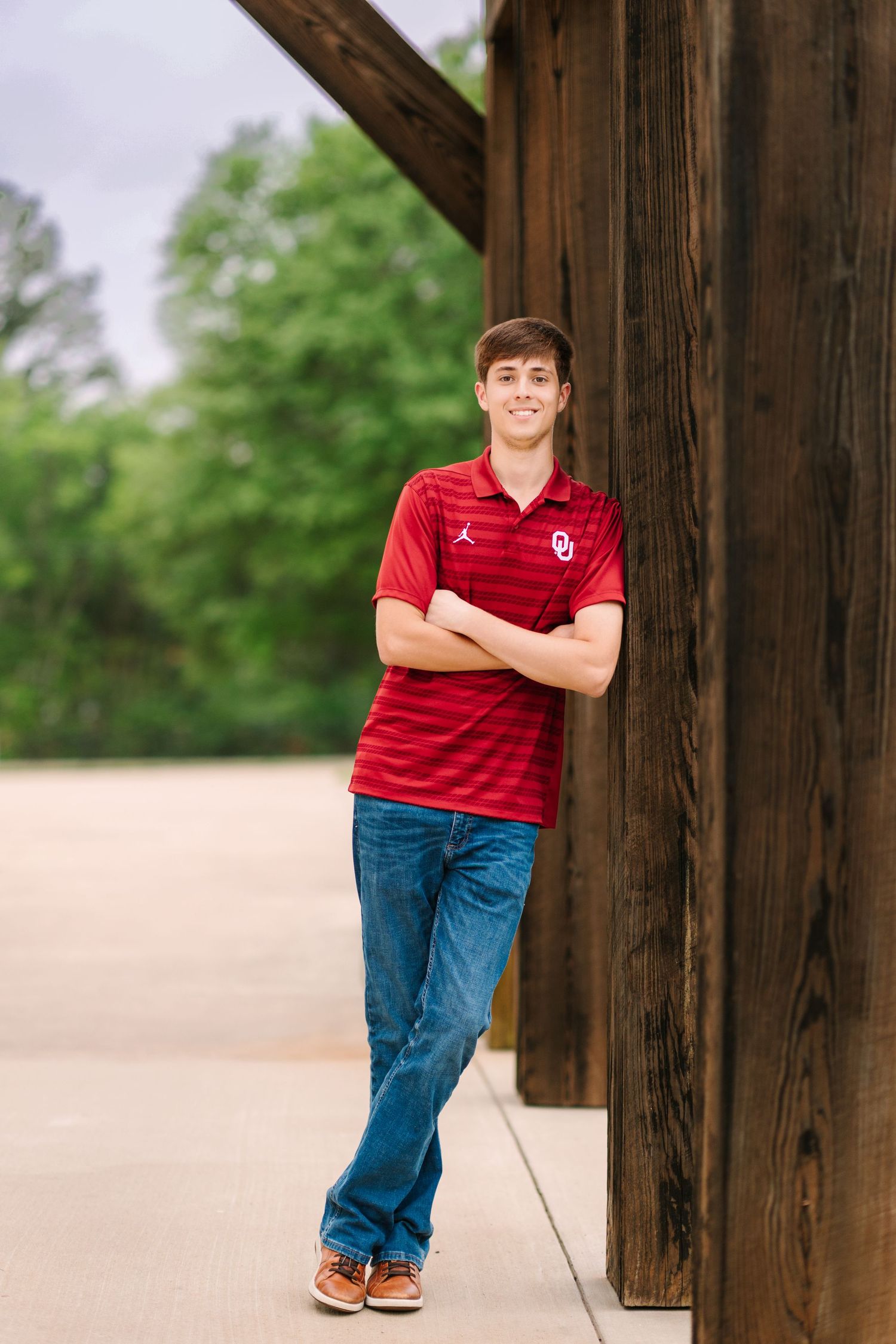 Student in red OU sports shirt and jeans leans against wooden post in casual outdoor portrait session.