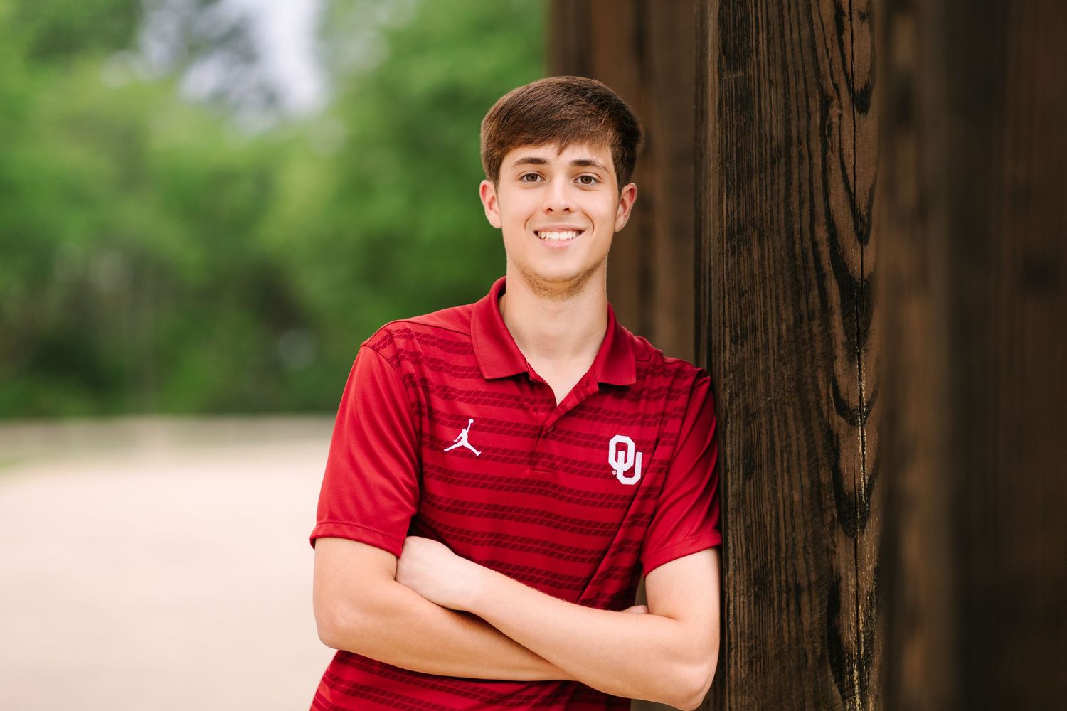Student in red Oklahoma Sooners polo shirt poses against outdoor wooded background in senior portrait.