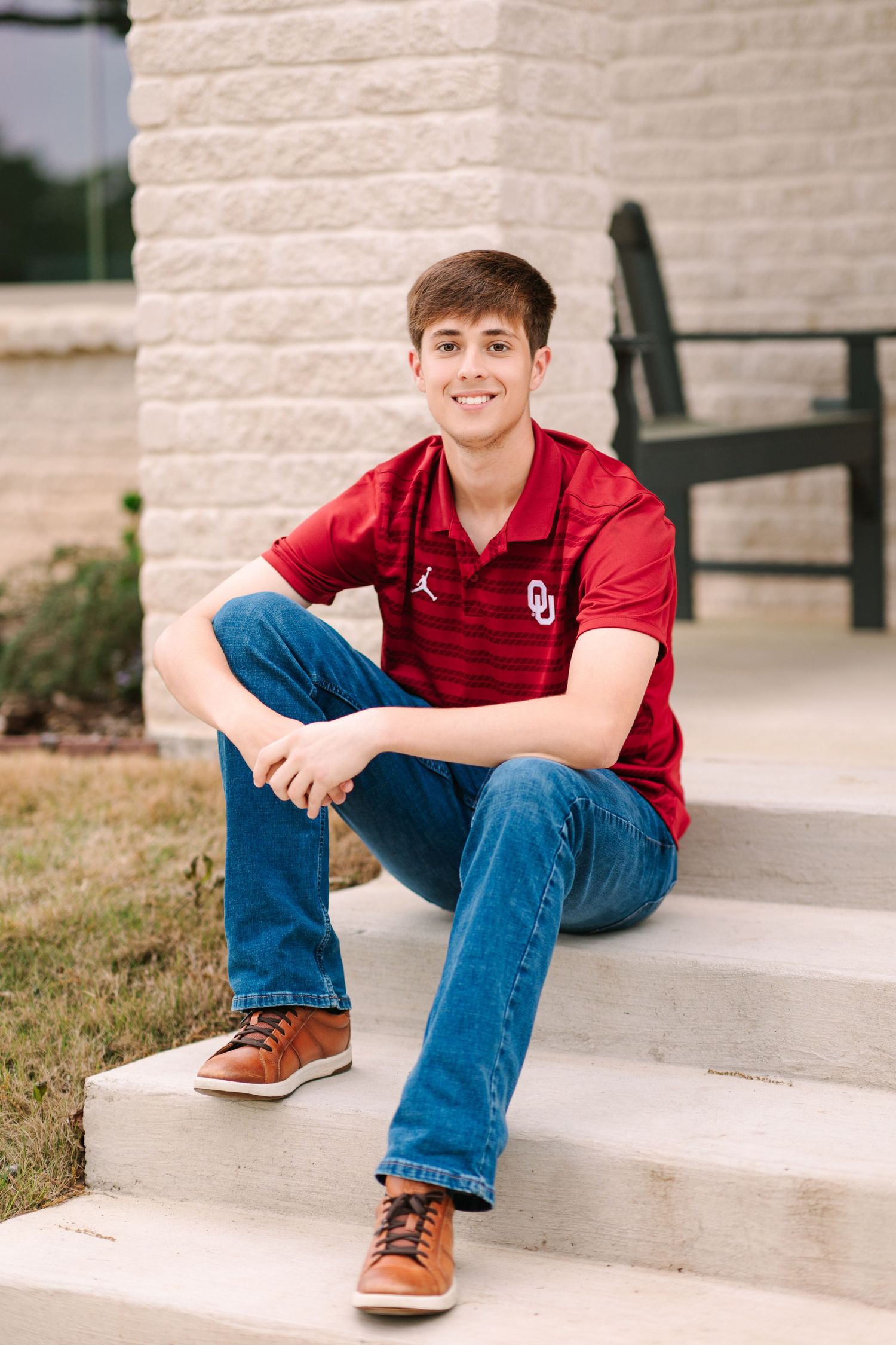 A person wearing a red Oklahoma shirt and blue jeans sits casually on concrete steps outdoors.