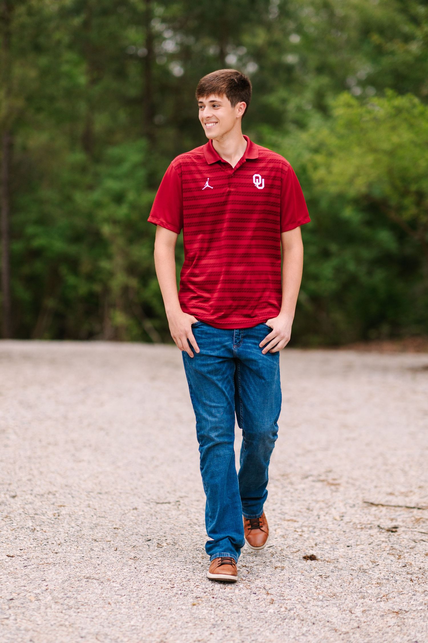 Student in crimson Oklahoma shirt and blue jeans walks across gravel surface against green background.
