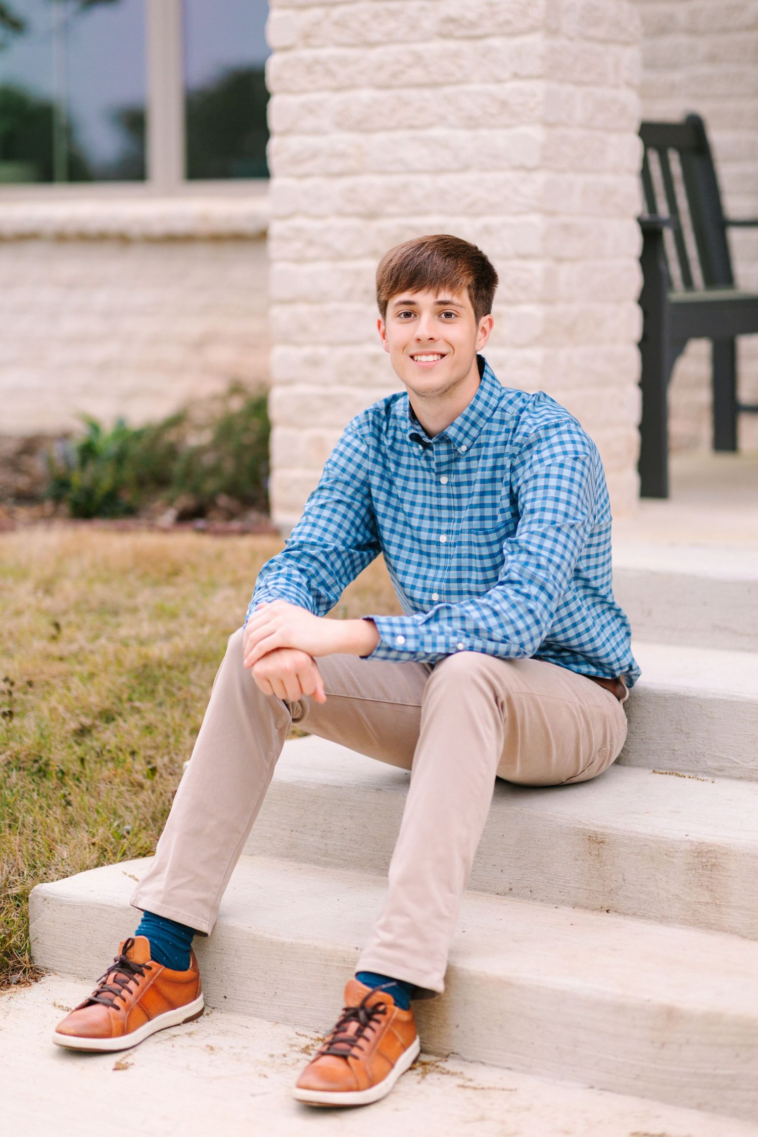 A person in a blue plaid shirt and khaki pants sits casually on concrete steps outdoors.