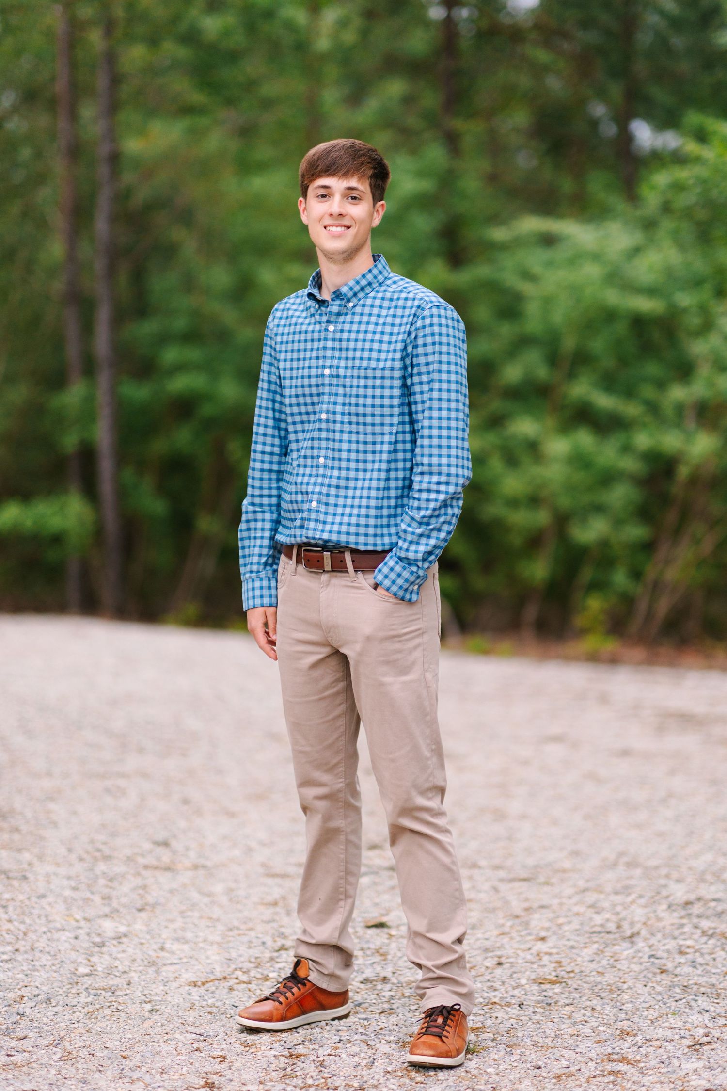 Full body view of someone in khaki pants and brown shoes standing on gravel path with trees in background.
