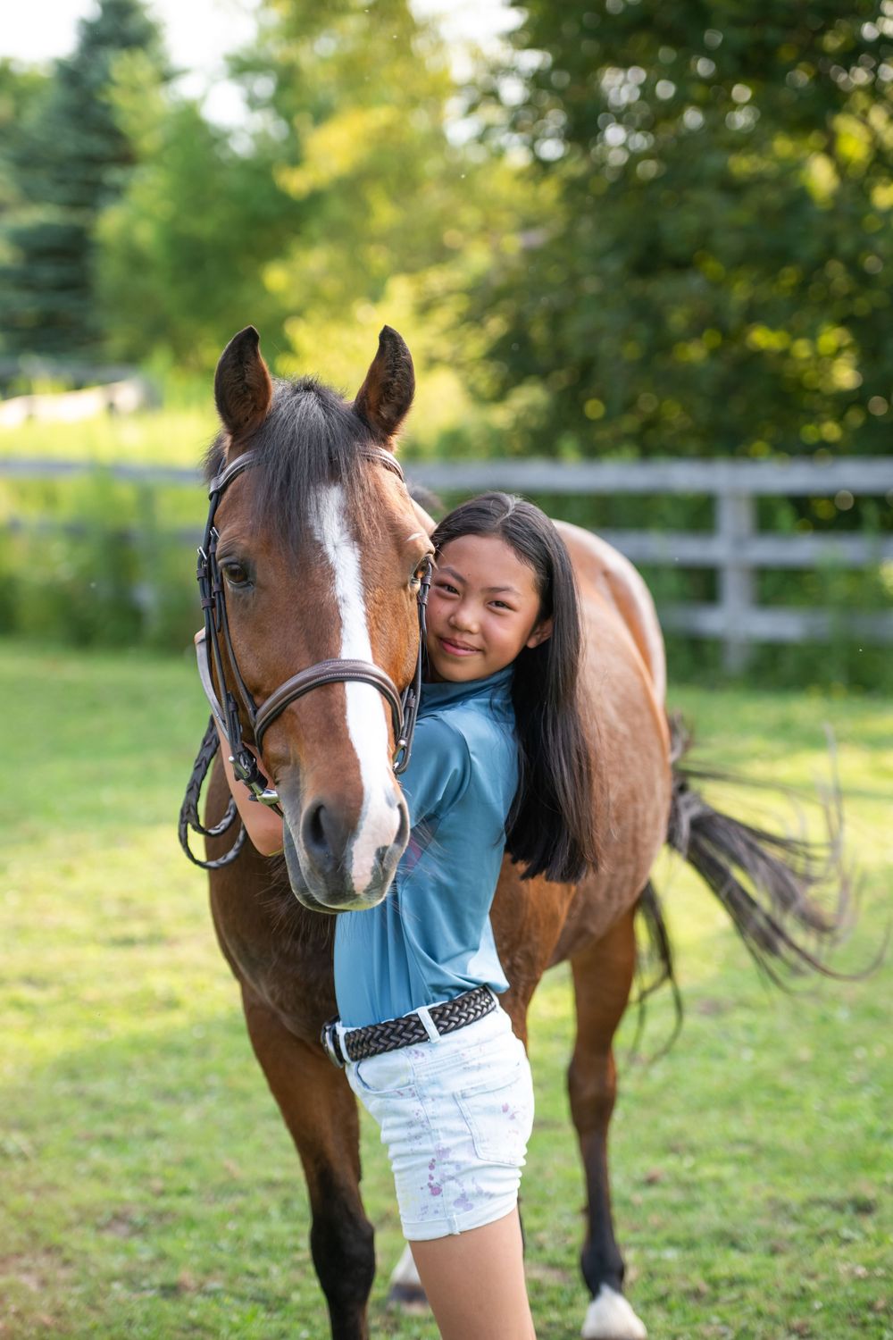 A brown horse with a white blaze is being hugged and affectionately embraced in an outdoor paddock with white fencing.