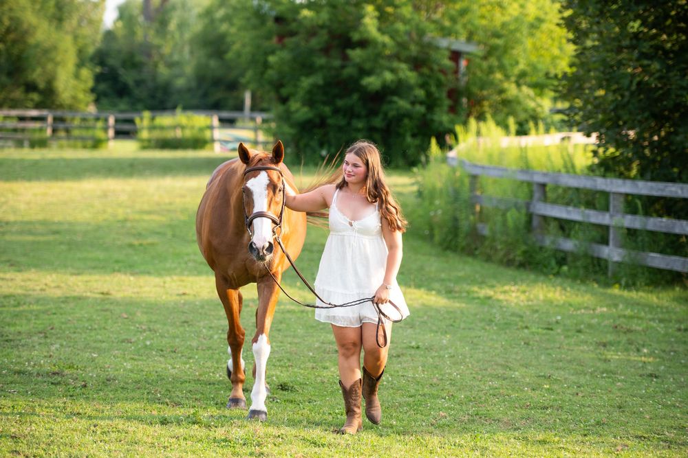 A person in white dress walking with a brown horse through a sunny green pasture along a wooden fence.