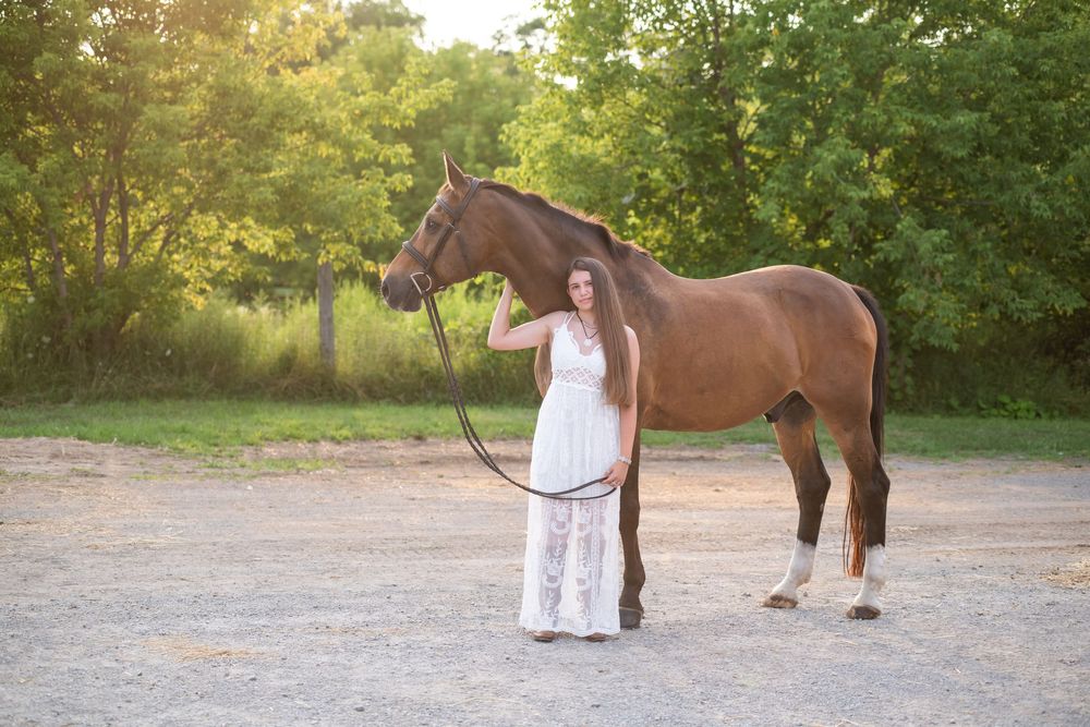 A bay horse walks alongside someone in white clothing through a sunlit wooded area during a photo session.
