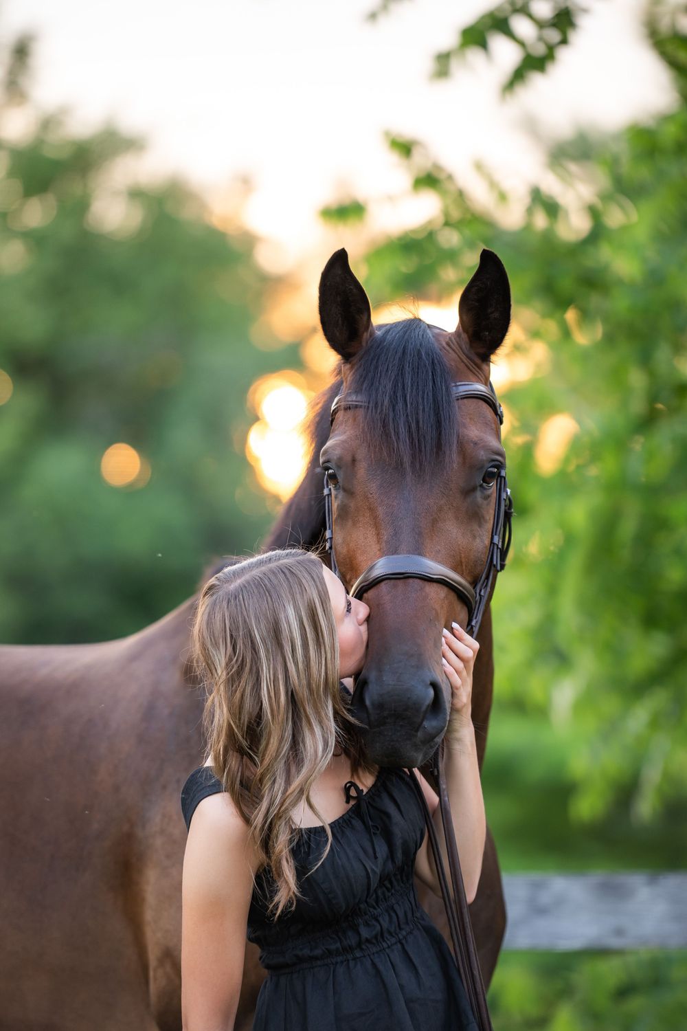 A woman in black stands next to a brown horse in front of lush green foliage at sunset.