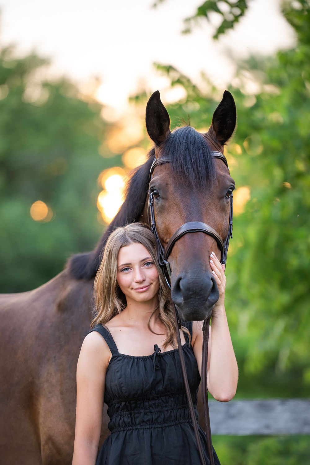 A beautiful brown horse poses alongside someone in a black dress in a warm sunset garden setting.