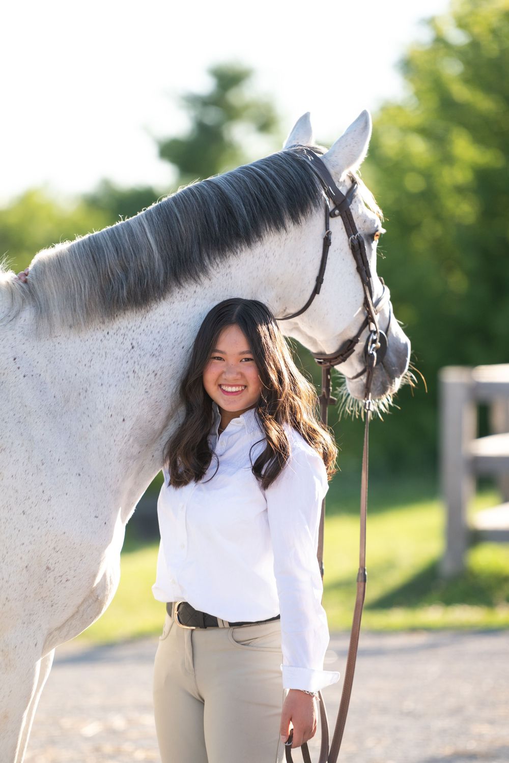 A rider in white shirt and beige pants stands next to a dappled grey horse in an outdoor arena setting.