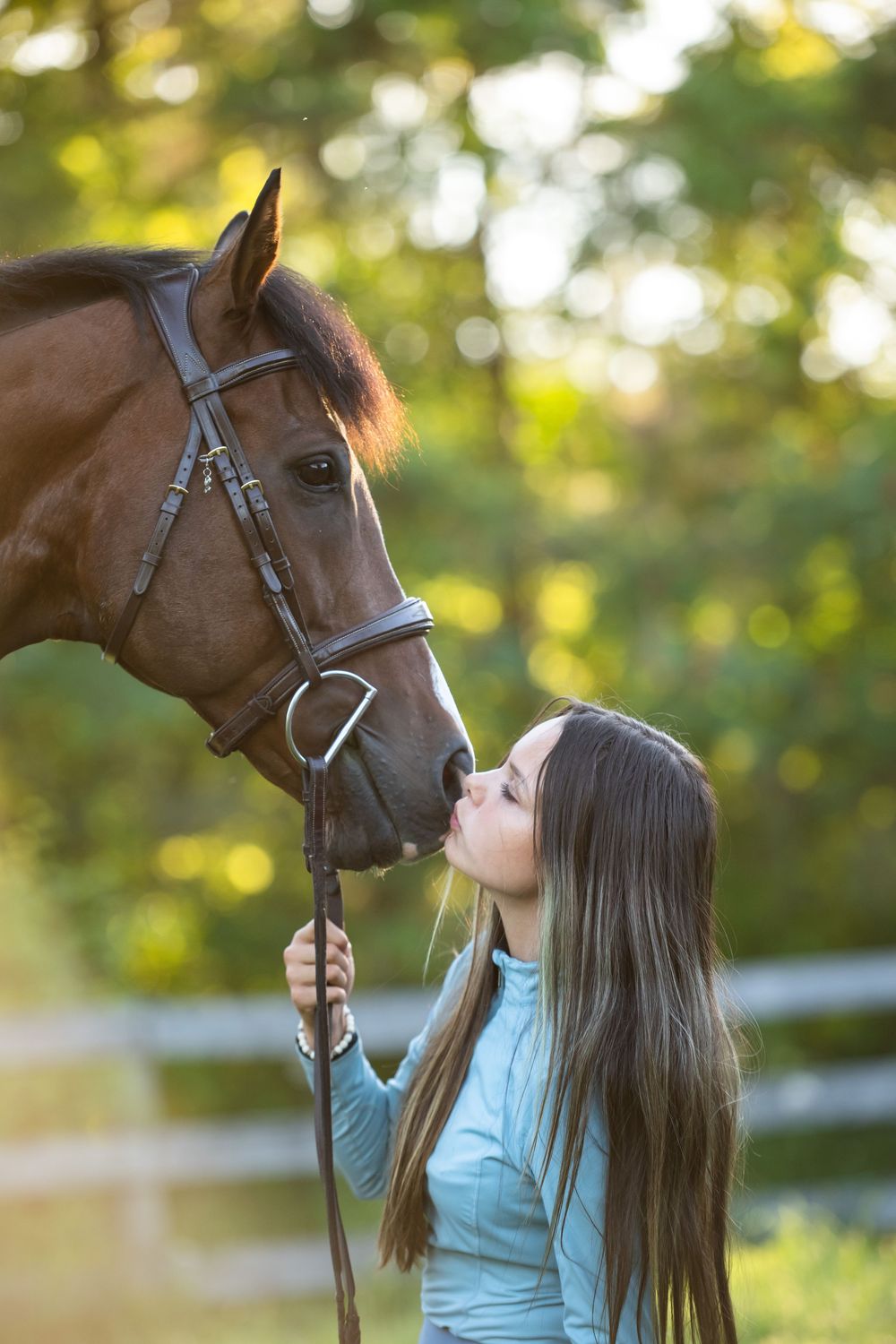 Tender moment between a rider in light blue shirt and a brown horse in a sun-dappled outdoor setting.
