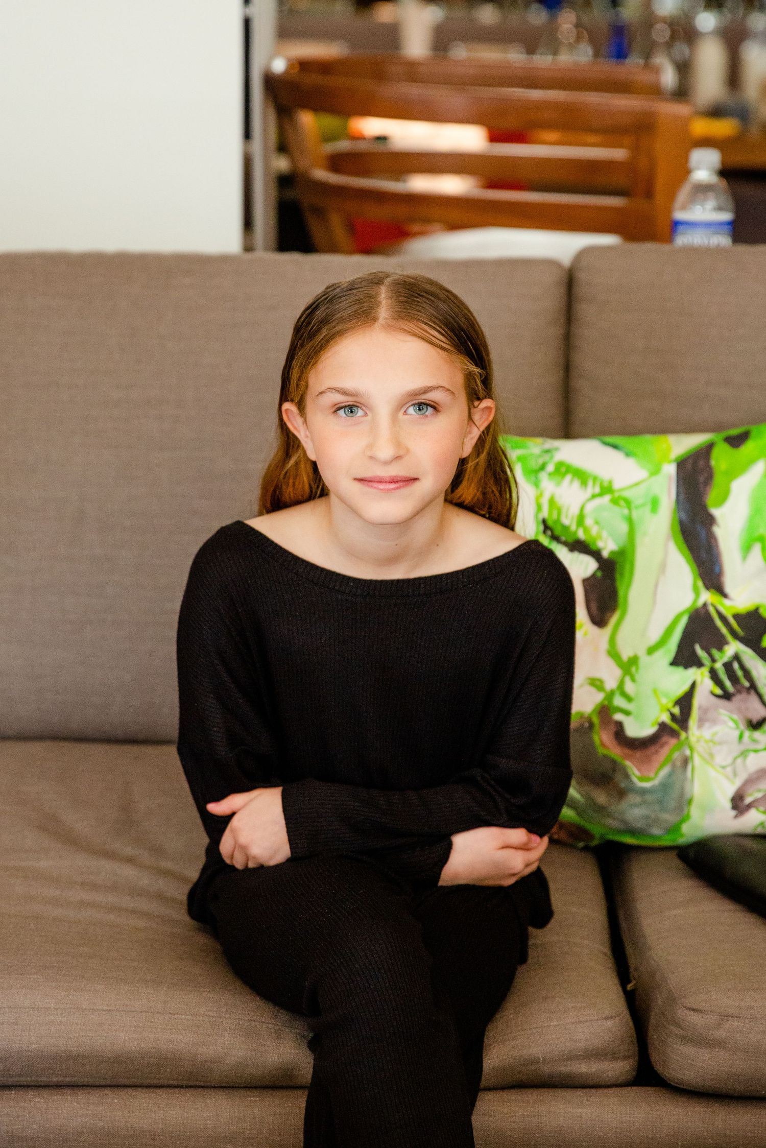 Young girl wearing black sitting on gray couch with decorative pillow in background.