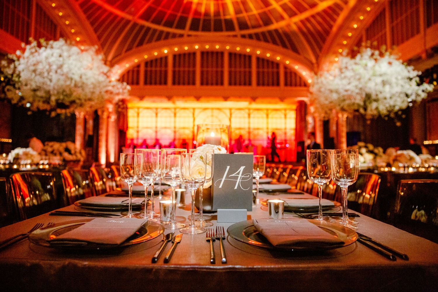 Elegant wedding reception table setting with table number and crystal glassware under dramatic red lighting.