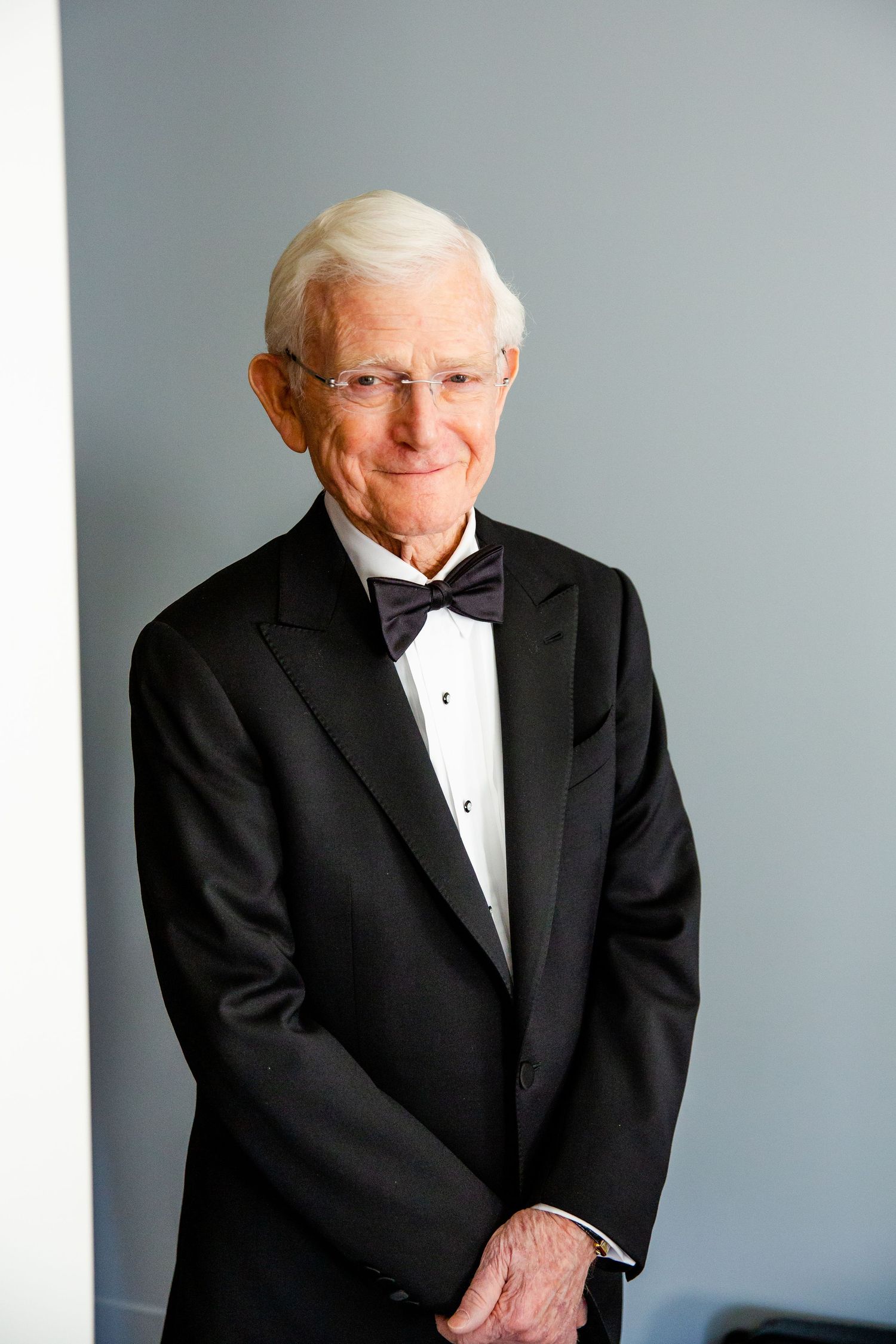 Formal portrait of elderly gentleman wearing black tuxedo and bow tie against gray background.