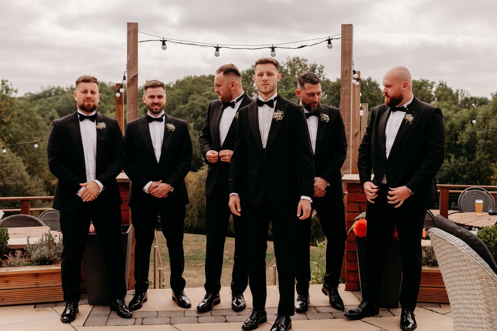 Group of wedding party groomsmen posing outdoors while wearing black tuxedos and laughing together.