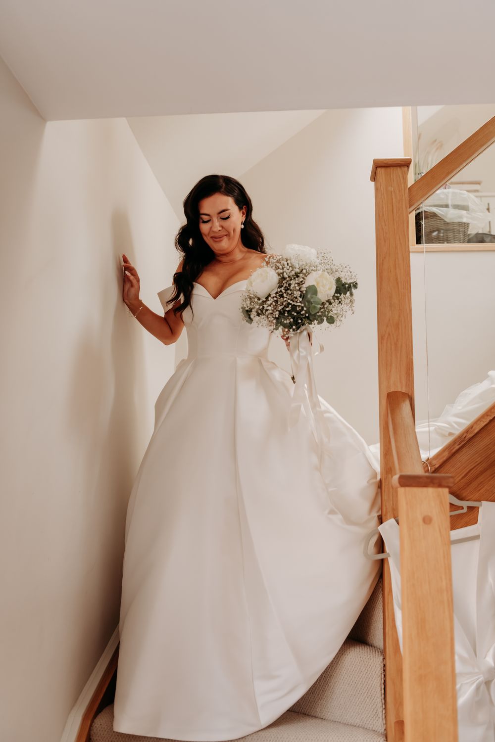 Beautiful bride in flowing white wedding gown gracefully ascends wooden staircase in intimate indoor setting.