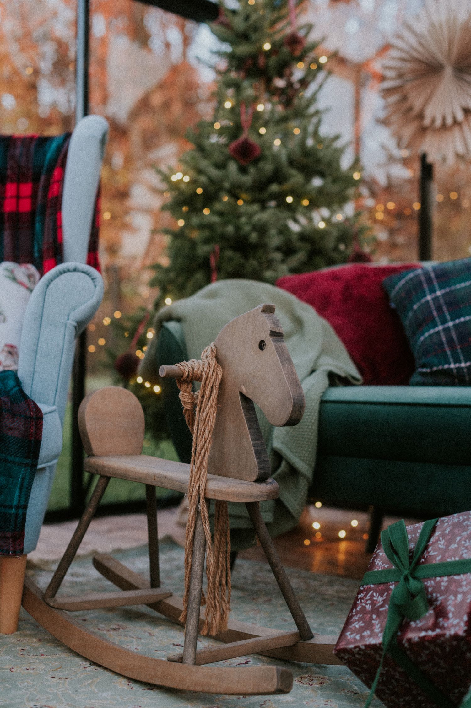 A cozy Christmas scene with a wooden rocking horse decoration near a decorated evergreen tree and plaid pillows.