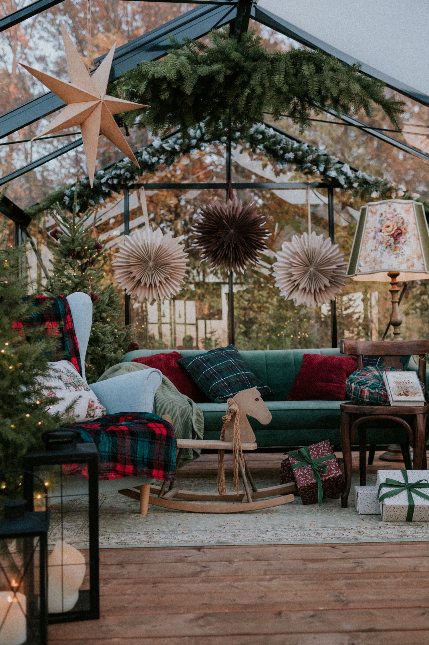 Cozy greenhouse decorated for Christmas with a teal couch, plaid pillows, and paper star decorations in a rustic setting.