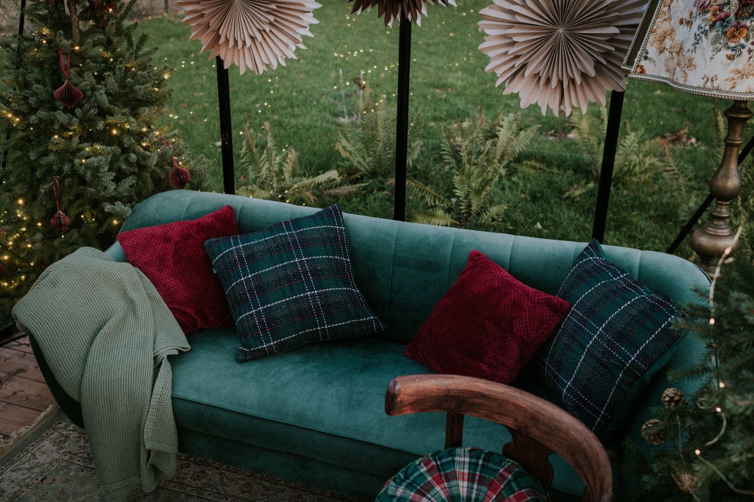 Close-up view of a teal velvet sofa with red and plaid throw pillows in a greenhouse setting.