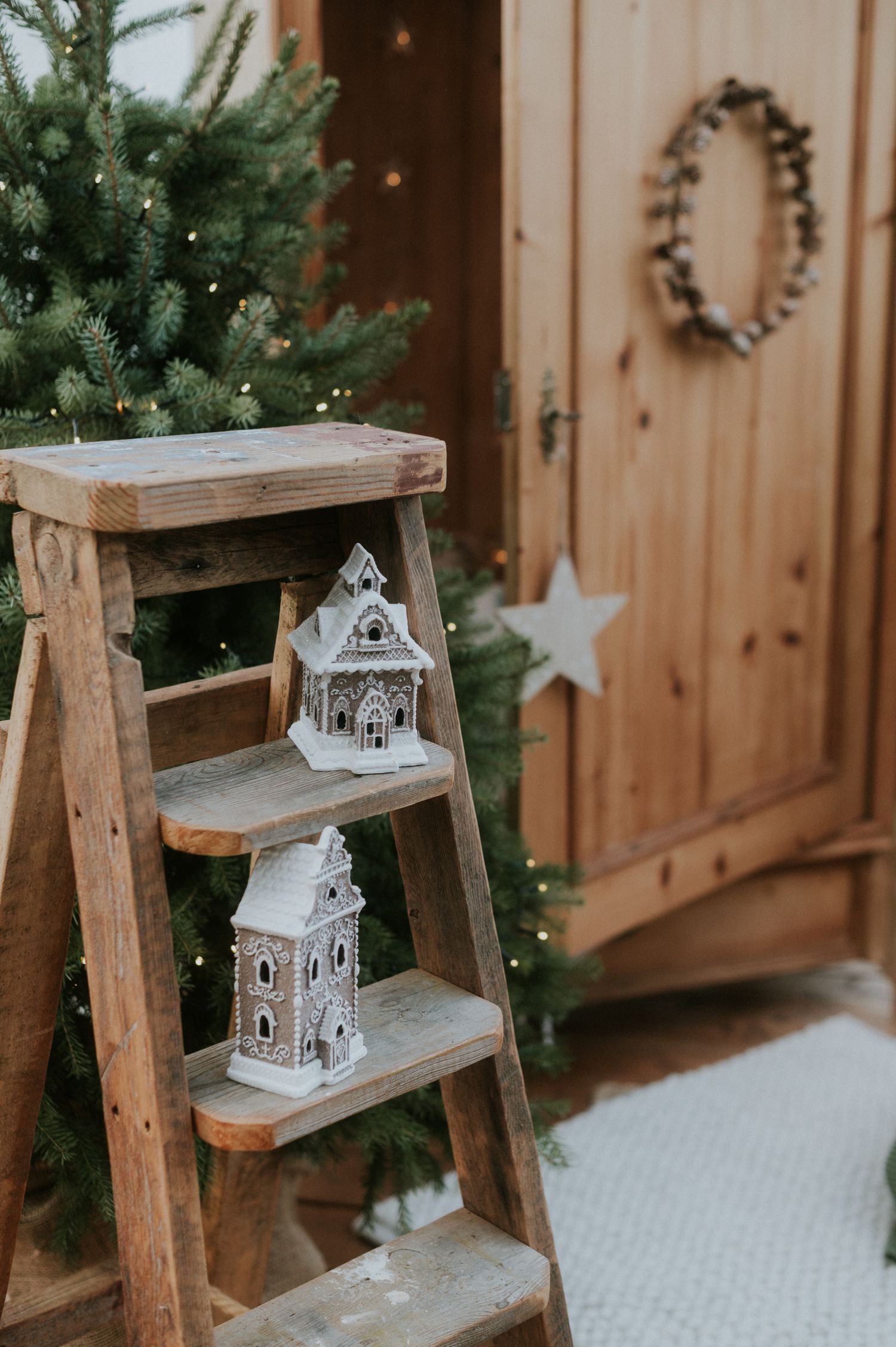 White ceramic Christmas houses displayed on wooden ladder steps beside evergreen tree and wooden door with wreath decoration.
