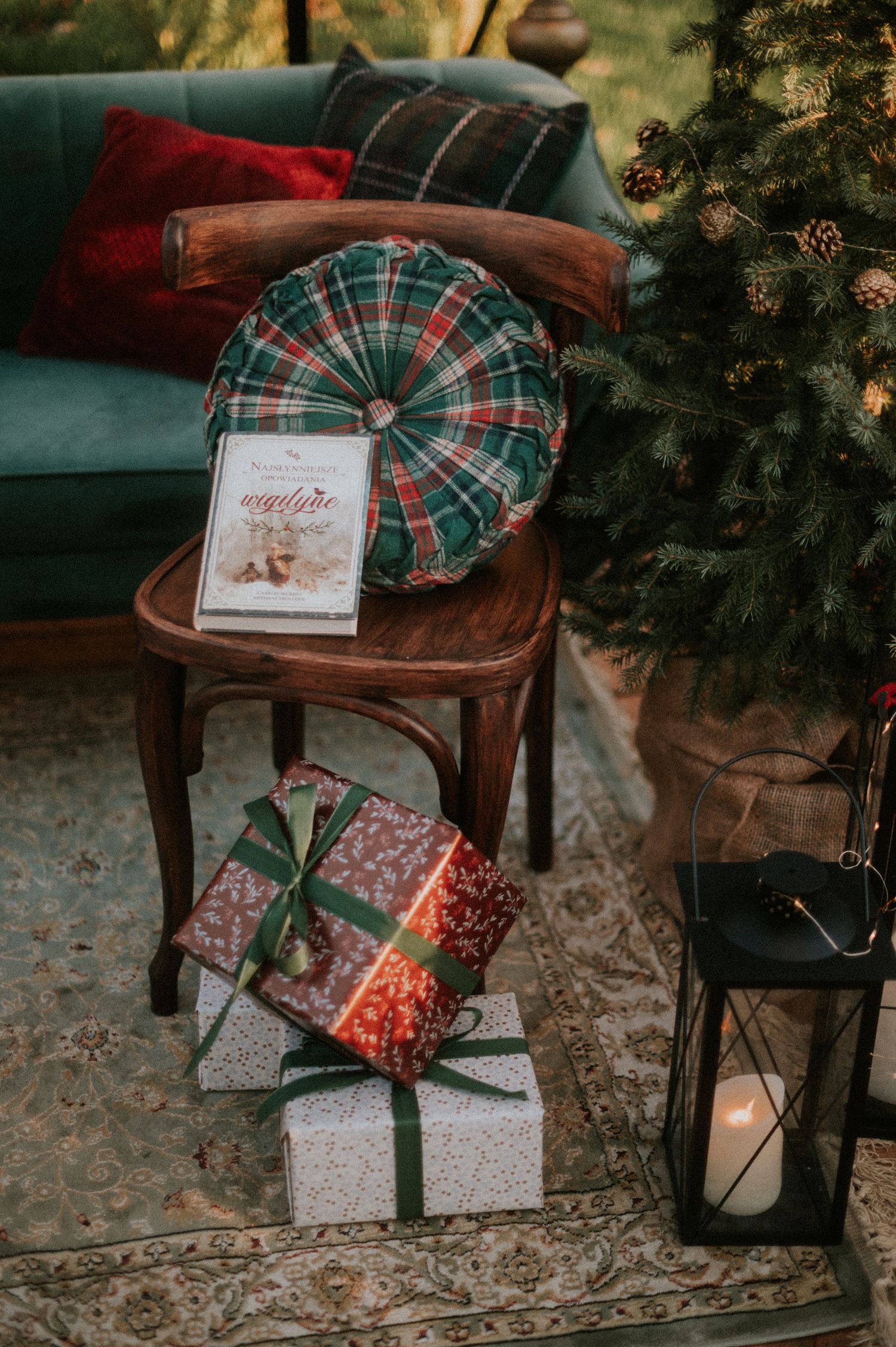 Christmas gifts wrapped in plaid and decorative paper sit beneath a rustic wooden chair next to a glowing lantern.
