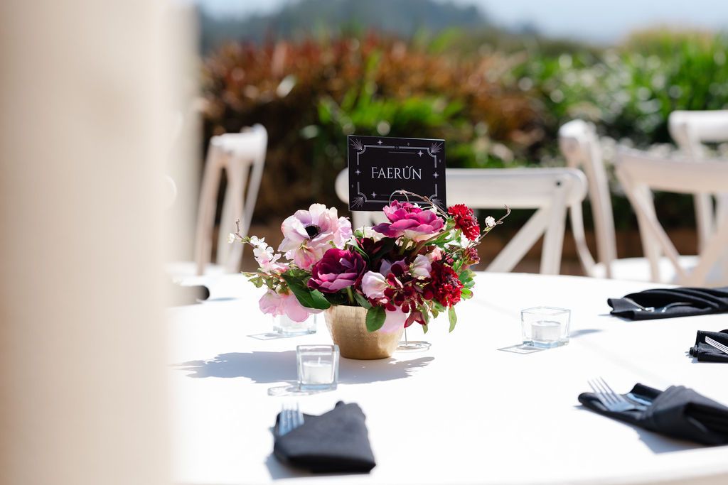 Pink and burgundy floral centerpiece with dark accents adorns an elegant table setting at an outdoor event.