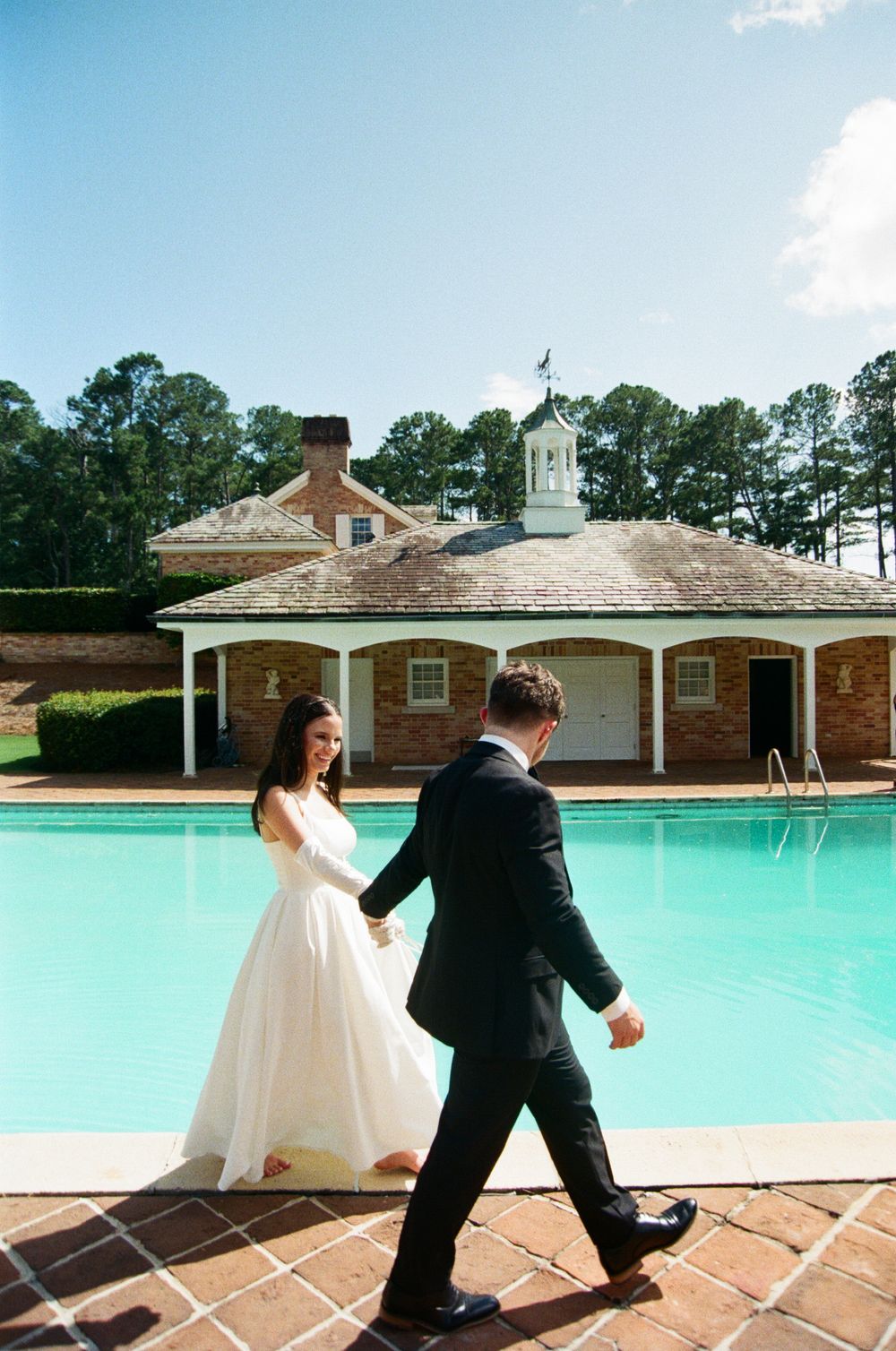 A couple in wedding attire playfully pose by a bright turquoise pool at an elegant brick venue.