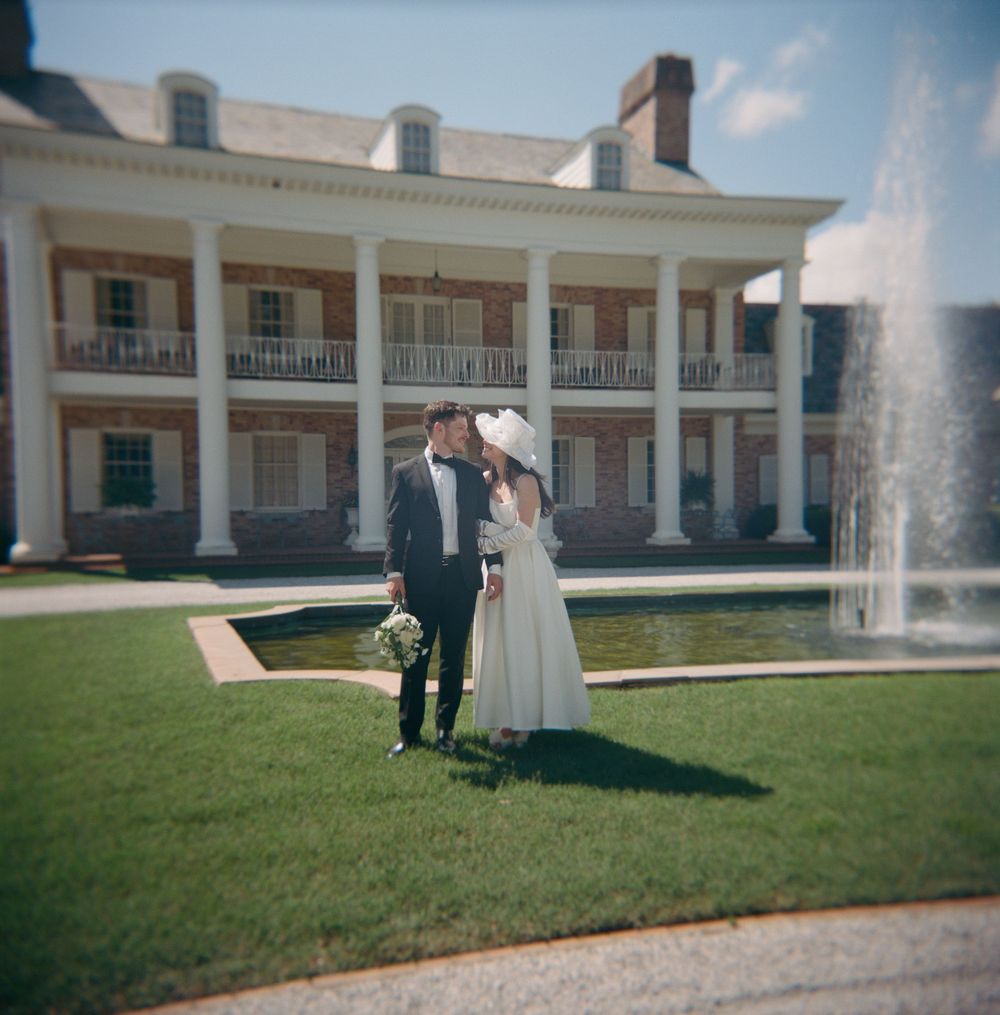 A romantic moment on a lawn in front of a grand southern mansion with white columns and a water fountain.