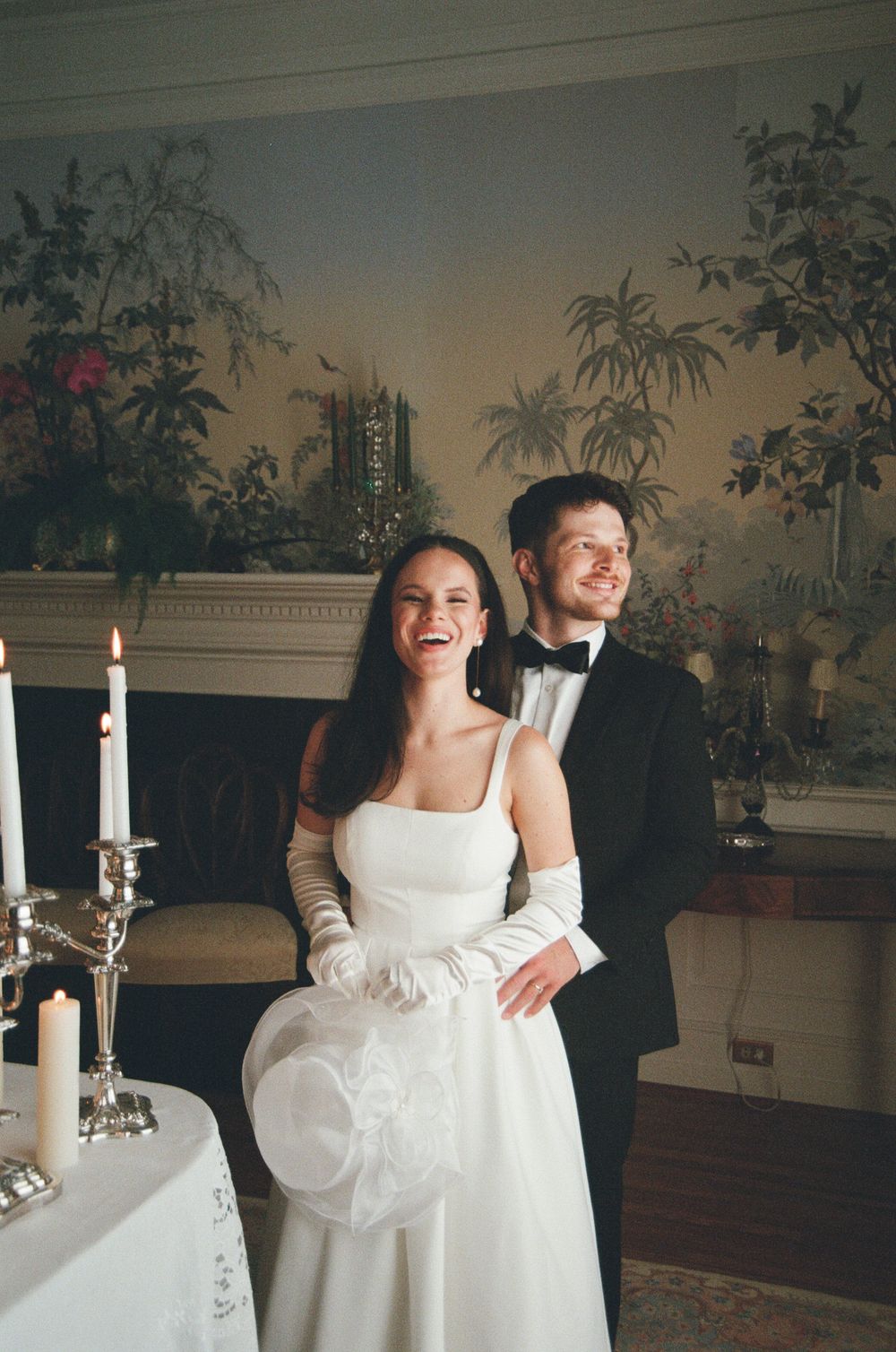 A couple shares a joyful moment at their wedding reception in an elegant room with vintage wallpaper and candlesticks.
