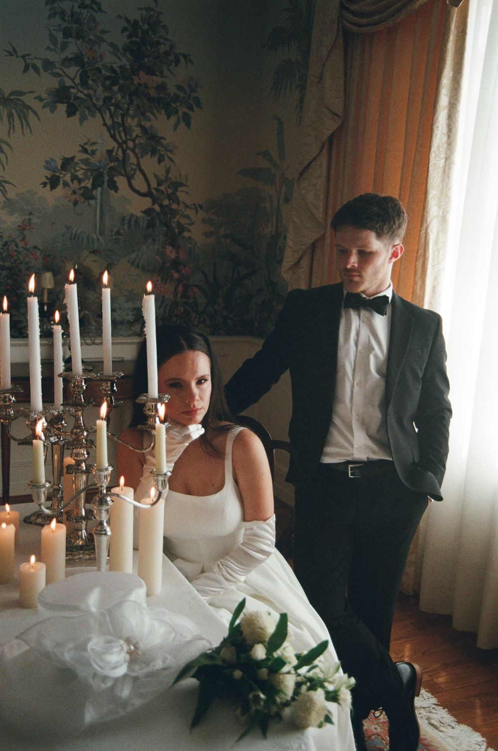 Elegant wedding scene with candelabras and white flowers in a room with vintage wallpaper and flowing curtains.