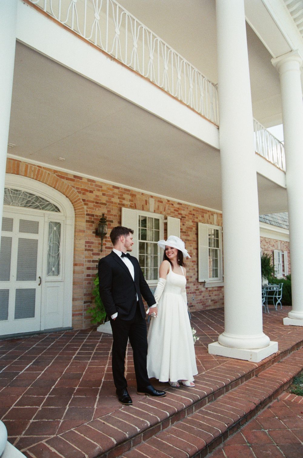 A couple in wedding attire poses on a brick porch in front of a grand southern-style mansion with white columns.