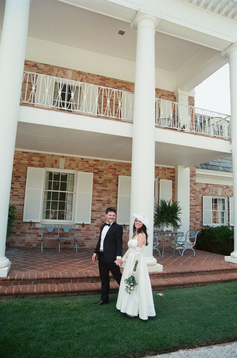 A formal couple stands on a manicured lawn in front of a two-story brick residence with white columns and balconies.