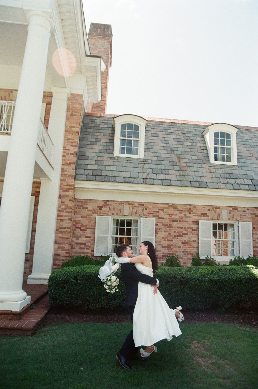 A couple in wedding attire dances playfully in front of a stately brick building with white columns on a sunny day.